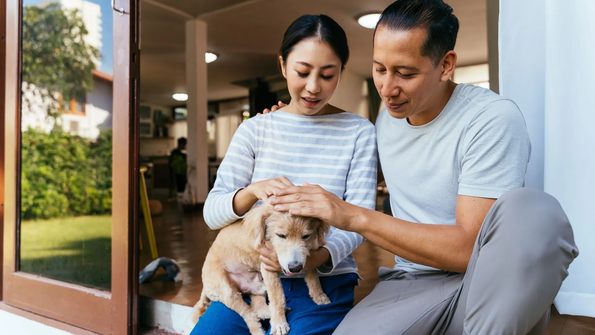 A couple sitting on their home’s porch, gently petting their small dog while enjoying a quiet moment together.