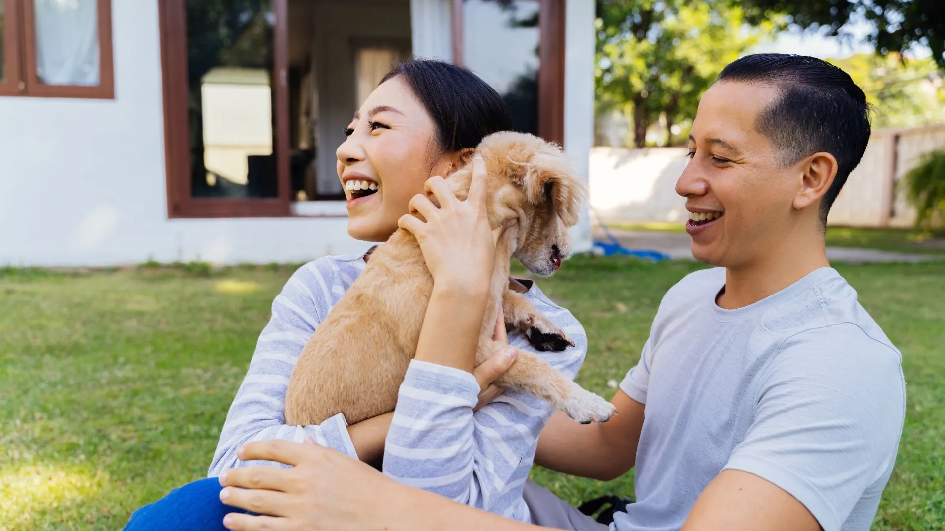 A smiling couple in their backyard, holding and hugging their small dog, expressing joy and affection.