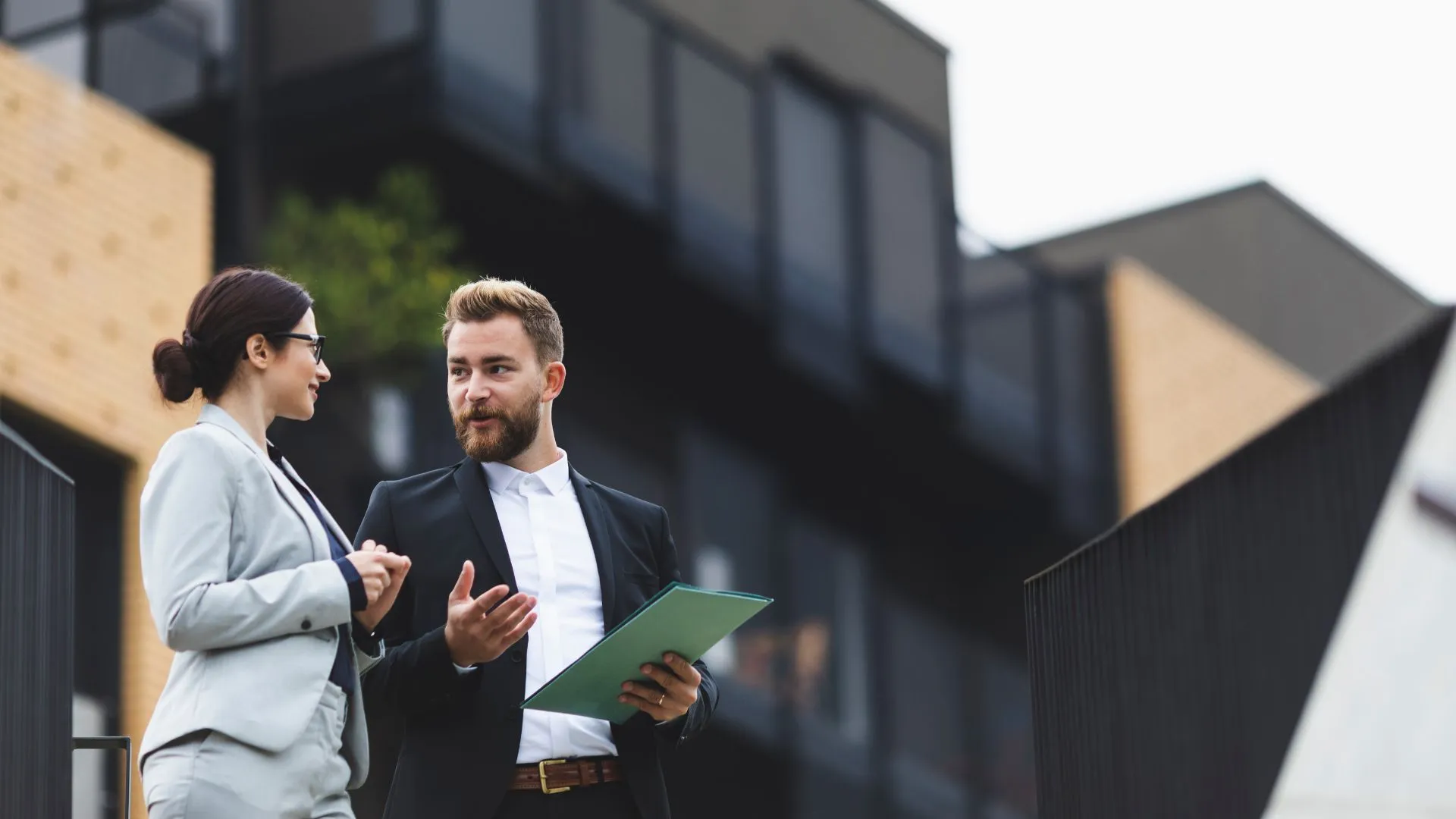 A professional man and woman in business attire discussing real estate opportunities in front of a modern building.
