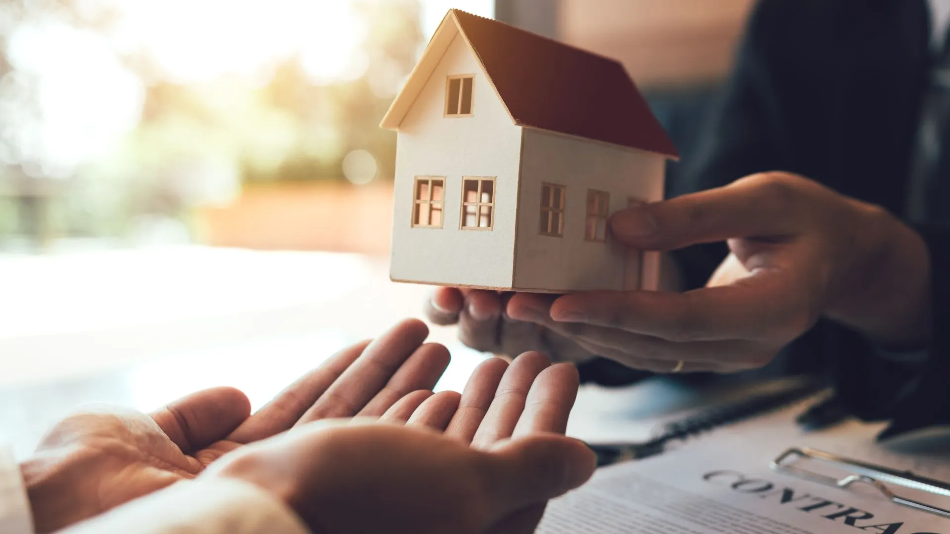 A symbolic image of a real estate transaction, with hands exchanging a miniature house model over a contract document.