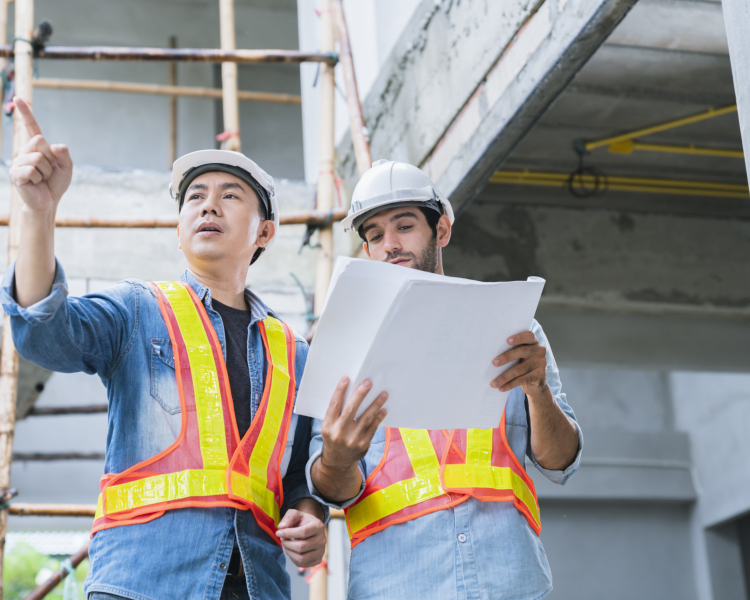 Young Asian engineers and caucasian engineer colleagues at site inspector and discuss together using notebook computer and paper blueprint at building construction site.home renovation ideas concept